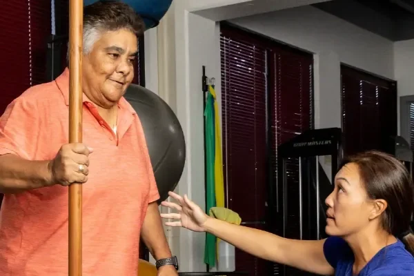 A patient standing with a wooden dowel for balance while a therapist talks him through the next movement